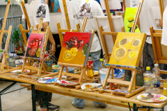 Auf einer Biertischgtarnitur stehen Tischstaffeleien, Pappteller als Paletten, Pinsel und Wassergläser. Auf den Staffeleien sind Kunstwerke in Acryl auf Leinwand der Schüler des deutsch-chinesischen Schüleraustausches in Neustadt an der Weinstraße zu sehen.